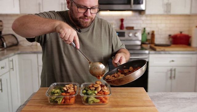 Man preparing healthy meal prep bowls filling containers with chicken, broccoli, carrots