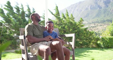 Laughing african american father and son relaxing on wooden bench in sunny mountain backyard