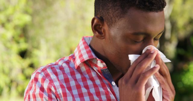 African American man sneezing into a tissue while outdoors. The young man is wearing a plaid shirt, suggesting a casual environment. Perfect for advertisements or articles related to healthcare, allergies, colds, flu, and seasonal changes.