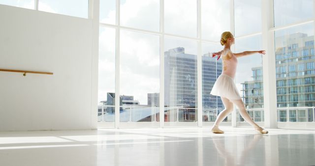 Young ballet dancer practicing in a bright, modern studio with tall windows that offer a view of city skyscrapers. Sunlight floods the room, enhancing the sense of grace and elegance. Useful for themes of ballet, urban life, performing arts, fitness, and inspirational contexts.