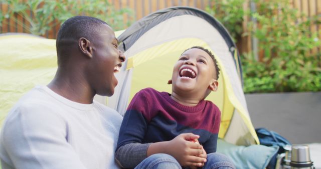 Father and son enjoying quality time together laughing outdoors near a tent. Ideal for use in family, parent-child bonding, camping, and summer activity themes. Perfect for marketing campaigns, blogs, advertisements promoting outdoor family fun, travel, and healthy relationships.