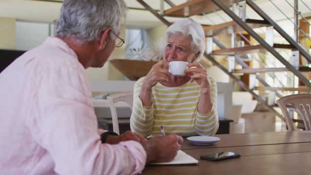 Senior Caucasian couple sitting at a wooden table, drinking coffee while discussing personal finances. The man is taking notes with a pen and paper. The woman is holding a cup, appearing thoughtful. Perfect for illustrating retirement planning, financial discussions, senior lifestyle, family finance decisions, or domestic life.