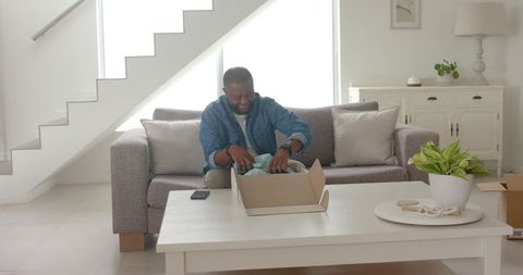 African american man unpacking new clothes on sofa in bright minimalist living room