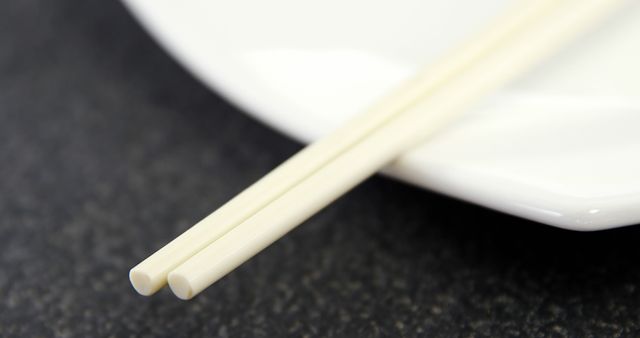 Detailed close-up of a pair of chopsticks resting on a white plate. Ideal for themes related to Japanese or Asian cuisine, minimalistic table settings, dining utensils, and food culture. Perfect for use in articles, blogs, and advertisements about dining, Asian food, or culinary arts.