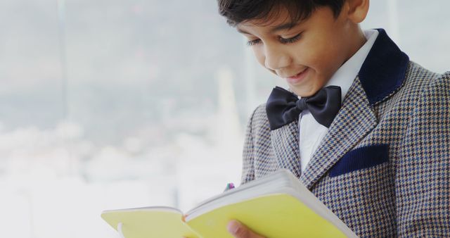 A young boy dressed smartly in a blazer and bow tie is engrossed in reading a book, with copy space. His focused expression suggests a love for learning or discovering a captivating story.