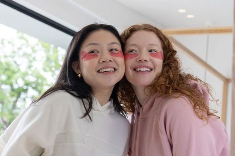 Two friends wearing pink under-eye gel patches smiling by window in cozy hoodies