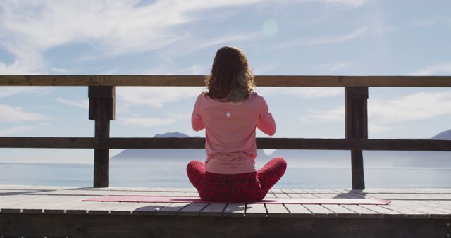 Woman sitting in a meditation pose on a wooden deck overlooking a calm ocean under a clear sky. The serene environment and natural setting make this perfect for usages involving relaxation, wellness retreats, yoga practice, mindfulness, or promoting a peaceful lifestyle. Ideal for wellness blogs, travel agencies, relaxation products, or meditation apps.
