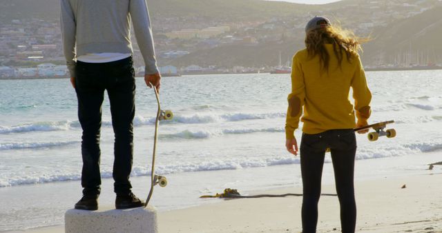 Two young individuals, in their teenage years, stand by the beach holding skateboards, with copy space. Their relaxed posture and the serene beach setting suggest a moment of leisure and enjoyment of the coastal environment.