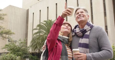 Senior couple exploring city holding coffee and pointing upward together in urban plaza