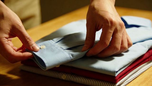Hands folding neat dress shirts on wooden table displaying buttons and layered fabrics