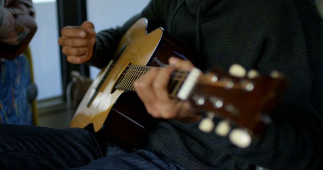 This close-up image focuses on a musician playing an acoustic guitar, with a detailed view of the hands and strings. Ideal for use on music-related websites, guitar lesson advertisements, or promotional materials for music classes, this image effectively showcases the intricacy and craftsmanship of playing a musical instrument. Perfect for illustrating musical creativity, practice, or performance.