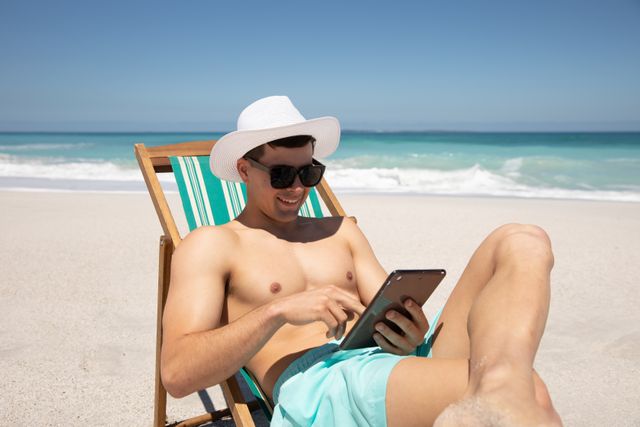 Man sitting in a deckchair on a sandy beach, wearing a sun hat and sunglasses, using a digital tablet. Ideal for promoting summer vacations, beach resorts, leisure activities, and digital lifestyle. Perfect for travel blogs, holiday brochures, and technology advertisements.
