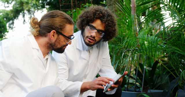 Two male botanists wearing lab coats and protective glasses reviewing information on a digital tablet in a greenhouse surrounded by various plants. This can be used to represent teamwork in scientific research, the integration of technology in botanical studies, or environmental science topics.