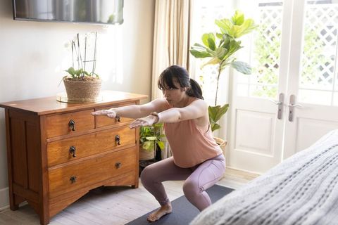 Asian woman performing squats on yoga mat in sunlit bedroom with indoor plants