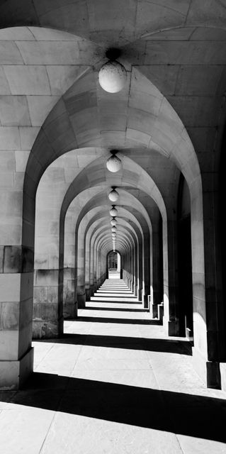 Monochrome view of a walkway with repeating arches creating a visually striking effect. The strong contrast between light and shadow highlights the architectural details. Ideal for use in projects related to architecture, design, and urban exploration or to add an artistic touch to interior design, advertising, and editorial pieces.