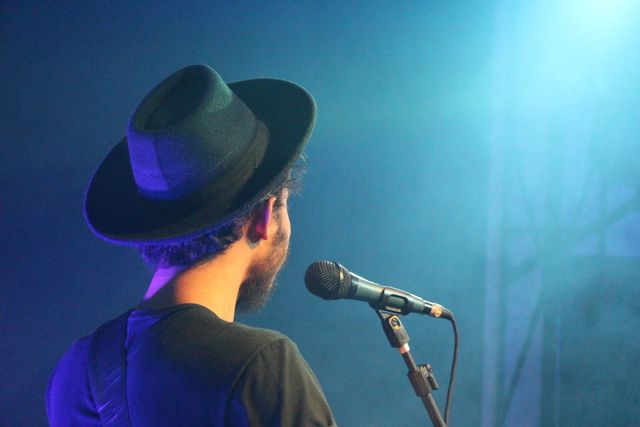 The artist is singing into a microphone on stage, illuminated by blue stage lights. Audience sees the back view of the performer, who is wearing a hat. Suitable for promoting live music events, concert tickets, nightlife venues, or artist brand promotions.