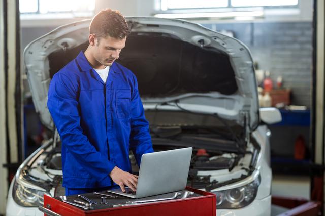Image shows a mechanic using a laptop, conducting diagnostics on a vehicle in an auto repair garage. The mechanic is wearing blue coveralls and working on a car with its hood open. This image can be used in automotive service contexts, repair shop advertisements, technology use in maintenance, and professional engineering services.