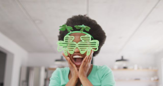 African American woman celebrating St. Patrick’s Day indoors, expressing excitement and joy while wearing green shamrock glasses. Perfect for advertisements, holiday greeting cards, cultural celebrations, and festive promotions.