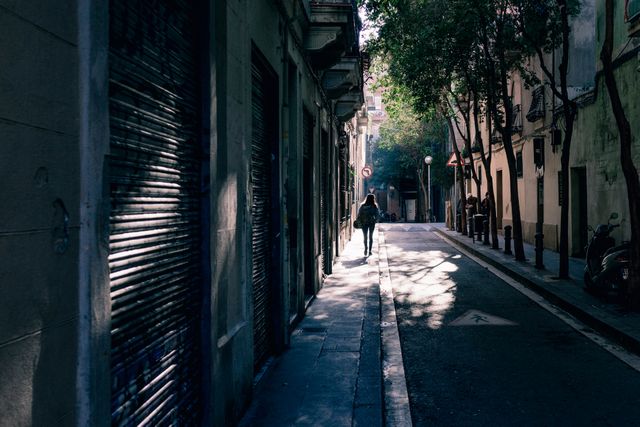 A lone pedestrian is silhouetted while walking down a deserted, sun-drenched urban street at dawn. The narrow street, lined with buildings casting long shadows on the sidewalk, evokes feelings of solitude and tranquility. Ideal for themes of urban life, solitude, early morning routine, and peaceful moments in the city.