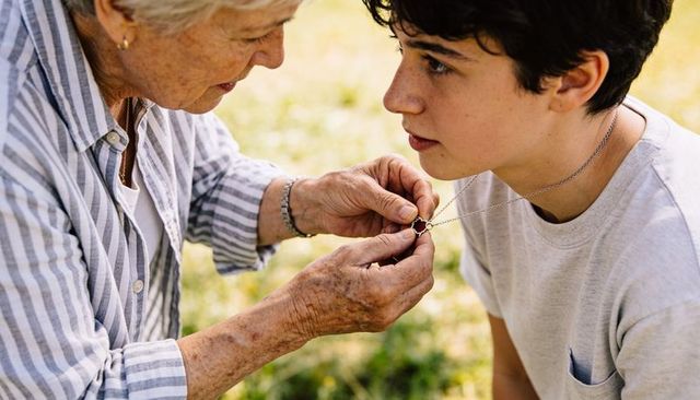 Elderly woman fastening necklace on teenage grandson outdoors, tender family moment, closeup
