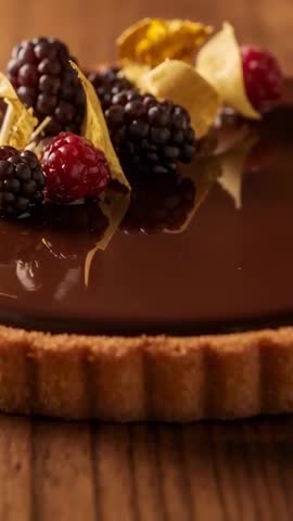 Vertical video panning over glossy chocolate ganache tart topped with berries and gold leaf