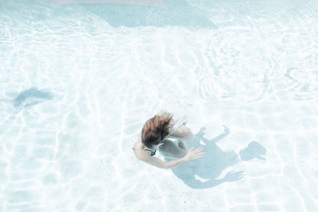 Woman swimming underwater with sunlight reflecting off the clear water. Perfect for promoting healthy lifestyle, aquatic fitness activities, swimwear advertisements, summer holiday graphics, relaxation concepts, and serene environmental imagery.