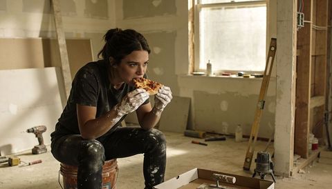 Female renovation worker eating pizza on upturned bucket during drywall remodeling break