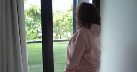 Contemplative african american woman standing at balcony door in pink hoodie looking outside