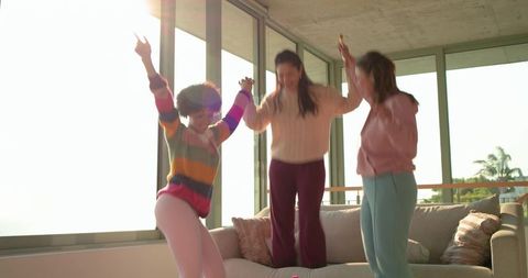 Sunlit diverse friends jumping on sofa, celebrating together in coastal living room