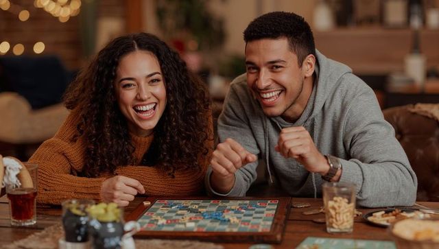 Couple laughing and playing board game at home, cozy warm evening with snacks and drinks