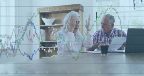Senior couple reviewing finances at home with laptop, paperwork and stock charts overlay