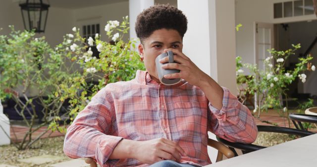Young man in a plaid shirt enjoying a cup of coffee on a patio. Ideal for themes of relaxation, leisure activities, morning routines, outdoor settings, casual lifestyle, and personal downtime. Suitable for advertising coffee brands, lifestyle blogs, or social media content about daily life.