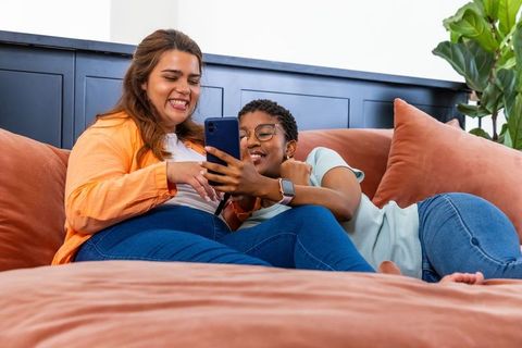 Two women laughing and sharing smartphone while relaxing on terracotta couch