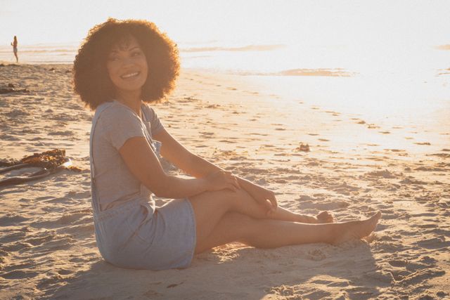 This image captures a joyful African American woman sitting on the beach during sunset, enjoying a moment of relaxation. Ideal for use in travel brochures, summer vacation promotions, wellness and lifestyle blogs, and advertisements focusing on leisure and relaxation.