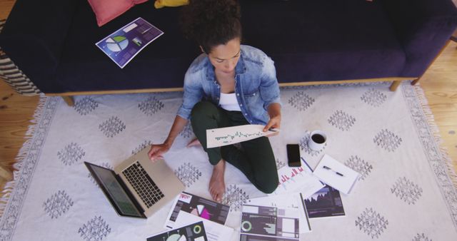 An overhead view of a young woman sitting on a rug, surrounded by data reports, working on her laptop. Coffee cup and papers spread around indicate intense focus and productivity in a casual living room setting. Ideal for concepts related to remote work, data analysis, business planning, multitasking, and modern work environment.