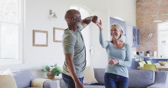 A mature couple smiling and dancing together in a cozy living room with modern decor. This represents a healthy and joyful lifestyle, showcasing love, happiness, and fun during retirement. Ideal for advertisements or content related to family life, relationships, retirement communities, health, and wellness.