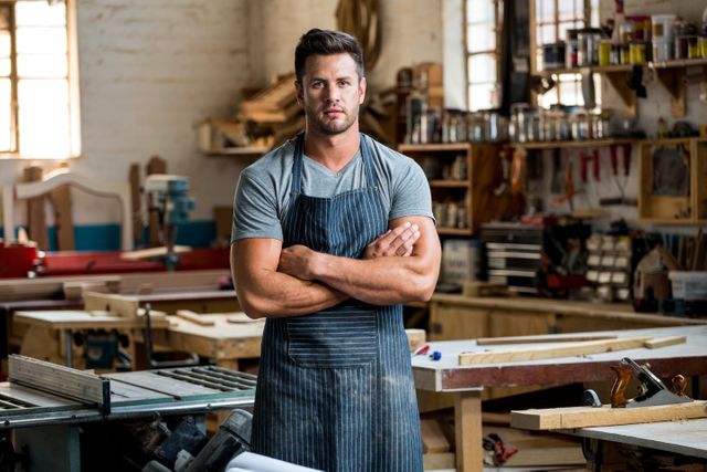 Carpenter standing with arms crossed in a dusty workshop filled with woodworking tools and materials. Ideal for use in articles or advertisements related to woodworking, craftsmanship, DIY projects, or industrial work environments.