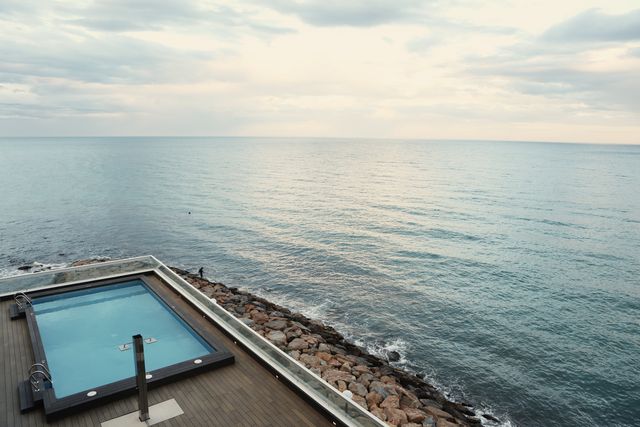 Beautiful infinity pool overlooking tranquil ocean during sunrise, perfect for promoting luxury vacations, seaside resorts, and peaceful retreats. The wooden deck and the expanse of calm waters add an element of elegance and serenity, ideal for travel brochures, wellness retreats, and relaxation-focused marketing materials.