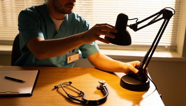 Healthcare worker adjusting desk lamp over wooden desk with stethoscope and id badge
