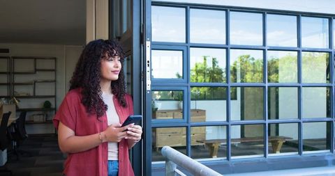 Young woman standing balcony holding smartphone looking out through glass office windows