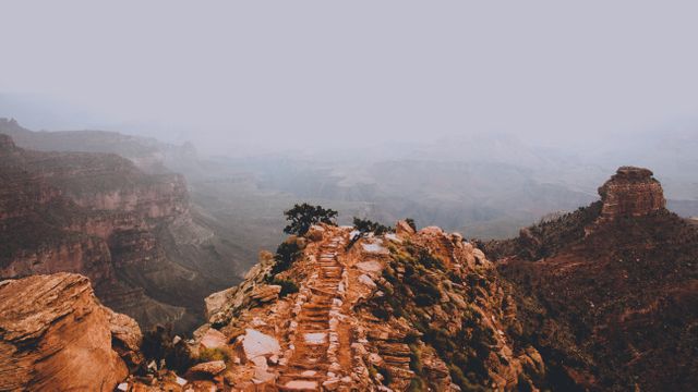 Rocky trail running through the Grand Canyon surrounded by mist offers breathtaking panoramic views suitable for adventure, hiking, and backpacking themes. Can be used in travel magazines, tourism ads, outdoor activity promotions, websites, or nature and photography blogs.