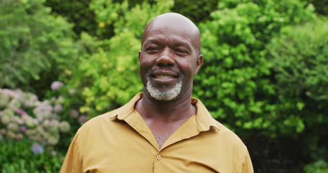 This image shows a smiling black man standing in a lush green garden. He is wearing a yellow shirt and exhibits a relaxed demeanor. Ideal for content focused on happiness, nature, outdoor activity, and lifestyle. Suitable for use in lifestyle blogs, gardening articles, and promotional materials highlighting outdoor relaxation and well-being.