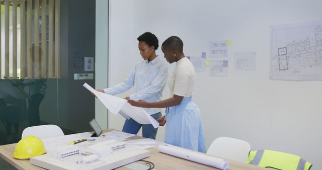 Two engineers discussing blueprints in a modern office. On the table are architectural plans, a hard hat, and other work materials. Ideal for images related to teamwork, professional collaboration, project management, engineering, and office environments.