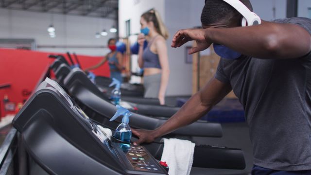 Man wearing face mask and headphones taking break while running on treadmill. Concept highlights fitness and health during a pandemic, showing adherence to safety measures such as mask-wearing and social distancing. Useful for topics on exercising safely during COVID-19, guidelines on gym precautions, health and wellness under quarantine.