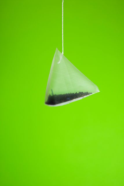 Pyramid tea bag hanging against bright green background creating a striking visual. Perfect for tea advertisement, health and wellness blogs, minimalist product displays, or packaging designs.