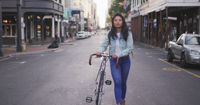 Young woman walking her bicycle down an empty city street. She is dressed casually in a denim jacket and blue jeans. This image can be used to convey themes of independence, urban lifestyle, and casual fashion. Ideal for use in advertisements, editorial content, and blogs discussing city living, personal journeys, or fashion.
