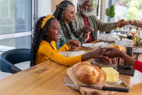 Multigenerational african american family sharing meal, breaking bread at sunlit wooden table