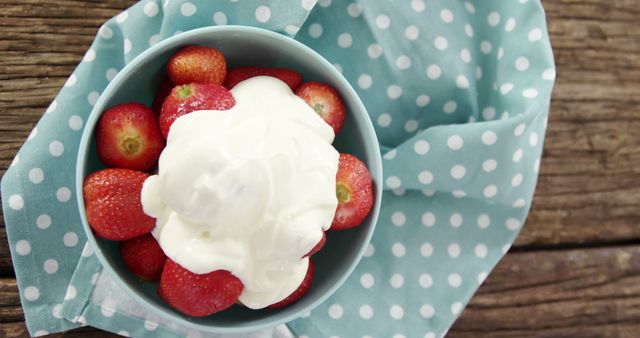 A bowl of fresh strawberries topped with a dollop of whipped cream sits on a wooden surface, with copy space. The vibrant red berries and creamy white topping create a tempting and delicious-looking dessert option.