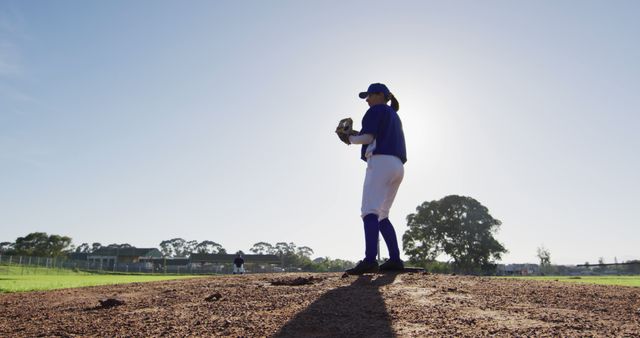 Youth baseball player standing on pitcher's mound under clear sky, captured from a low angle. Perfect for use in sports-related promotions, youth athletic program advertisements, outdoor activities campaigns, and similar themed projects regarding teamwork, focus, and dedication.