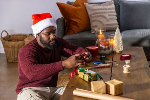 African american man wearing santa hat wrapping christmas gifts at cozy living room table